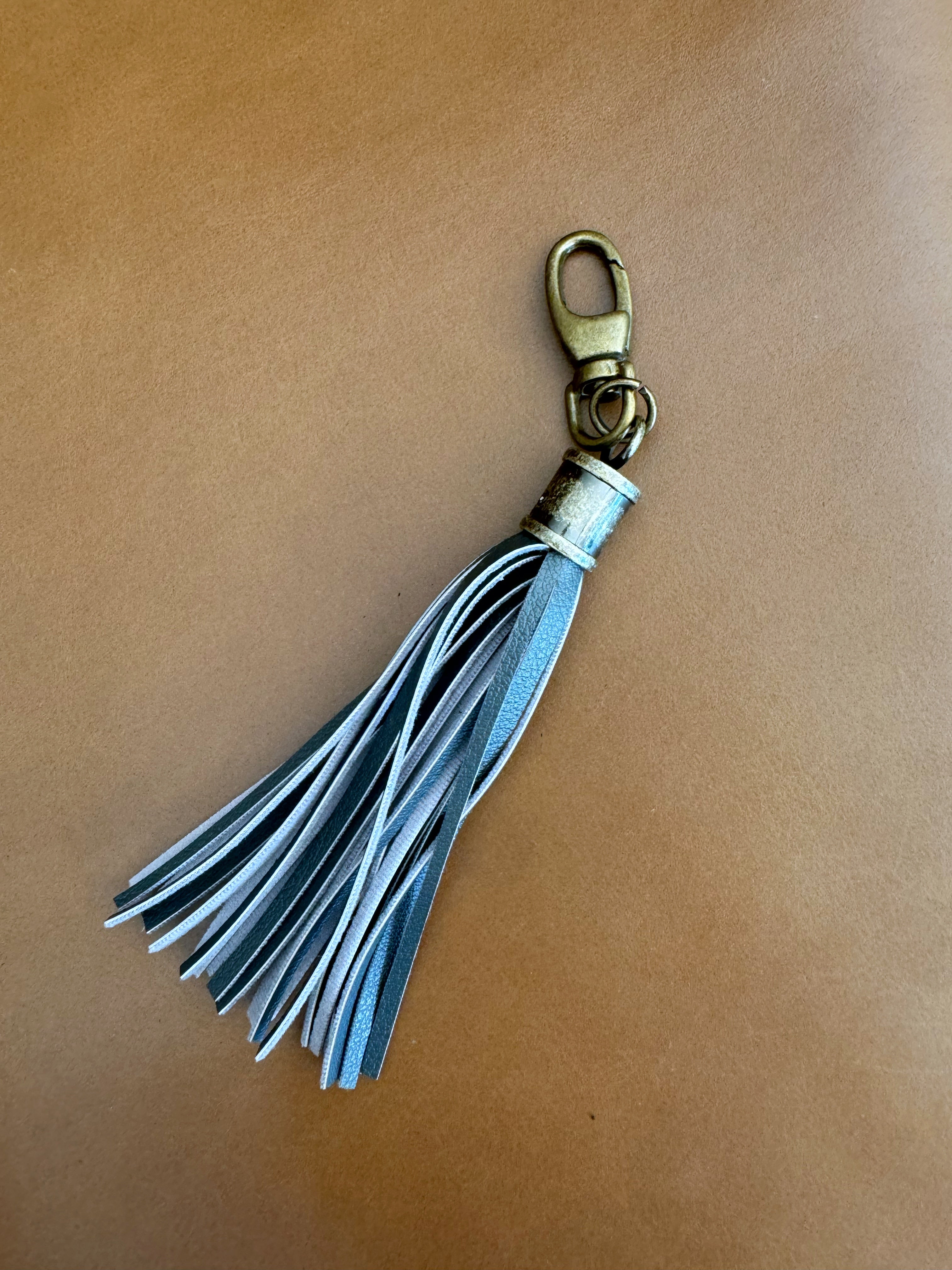 Tassel with thin gray vinyl fringe and cylindrical antique brass Hardware. Lobster clasp easily attaches to bags and accessories.