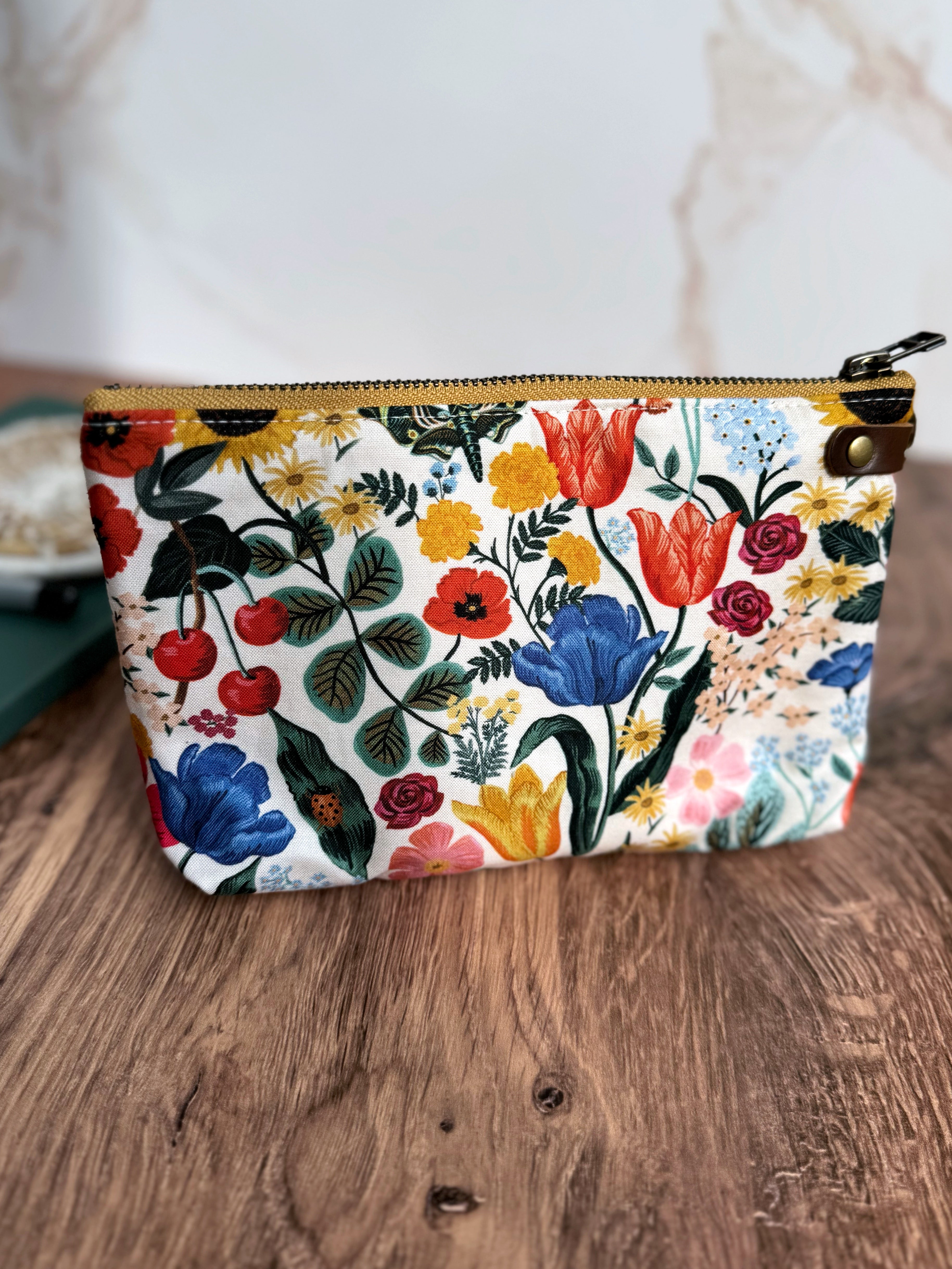 Rifle Paper Co. fabric pouch, white background and bright colors and a yellow zipper and leather tab on the side.