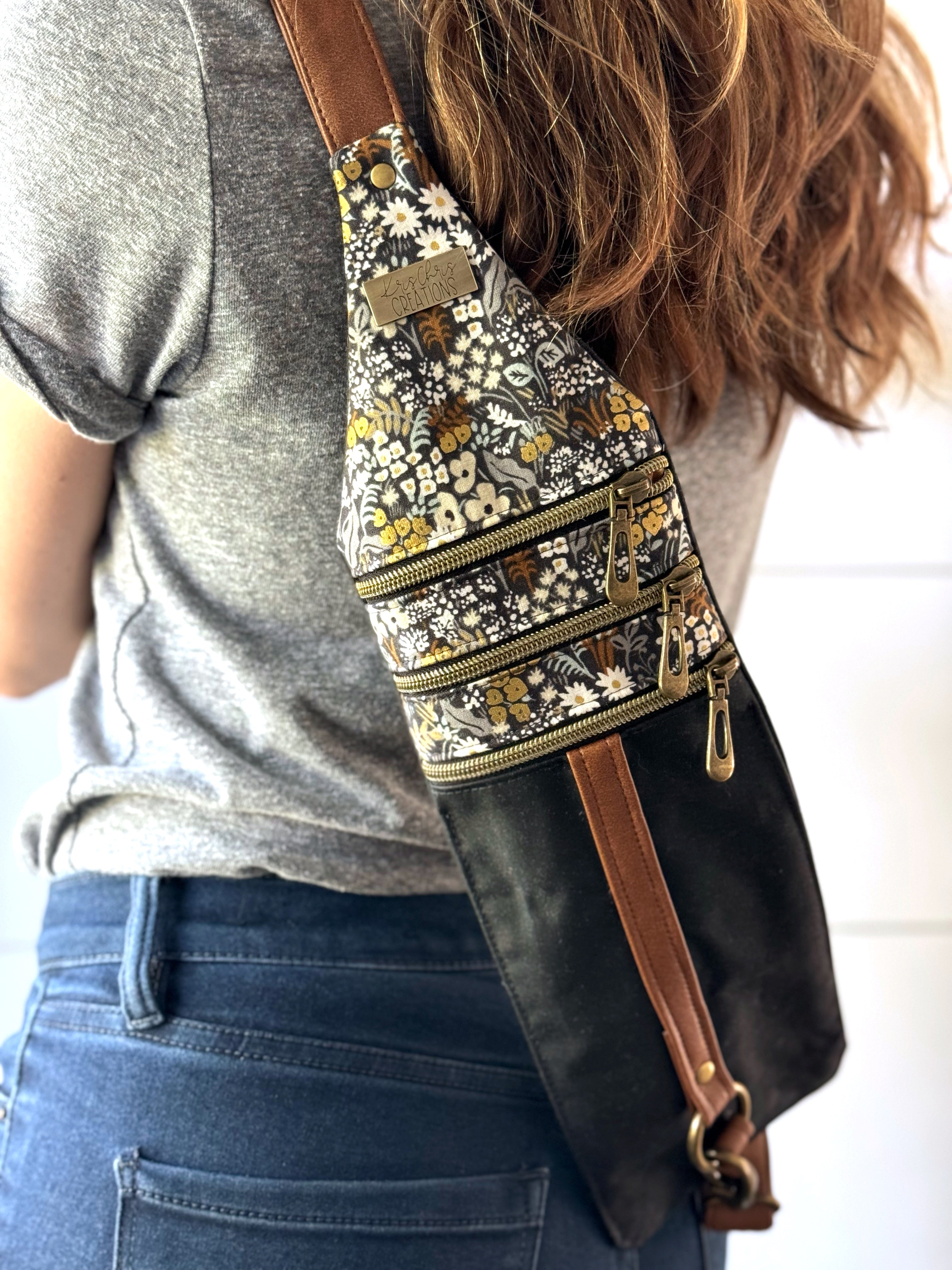 Woman in jeans wearing black and tan floral crossbody bag on the back. Showing the 3 brass zippers across the top and adjustable vinyl strap.