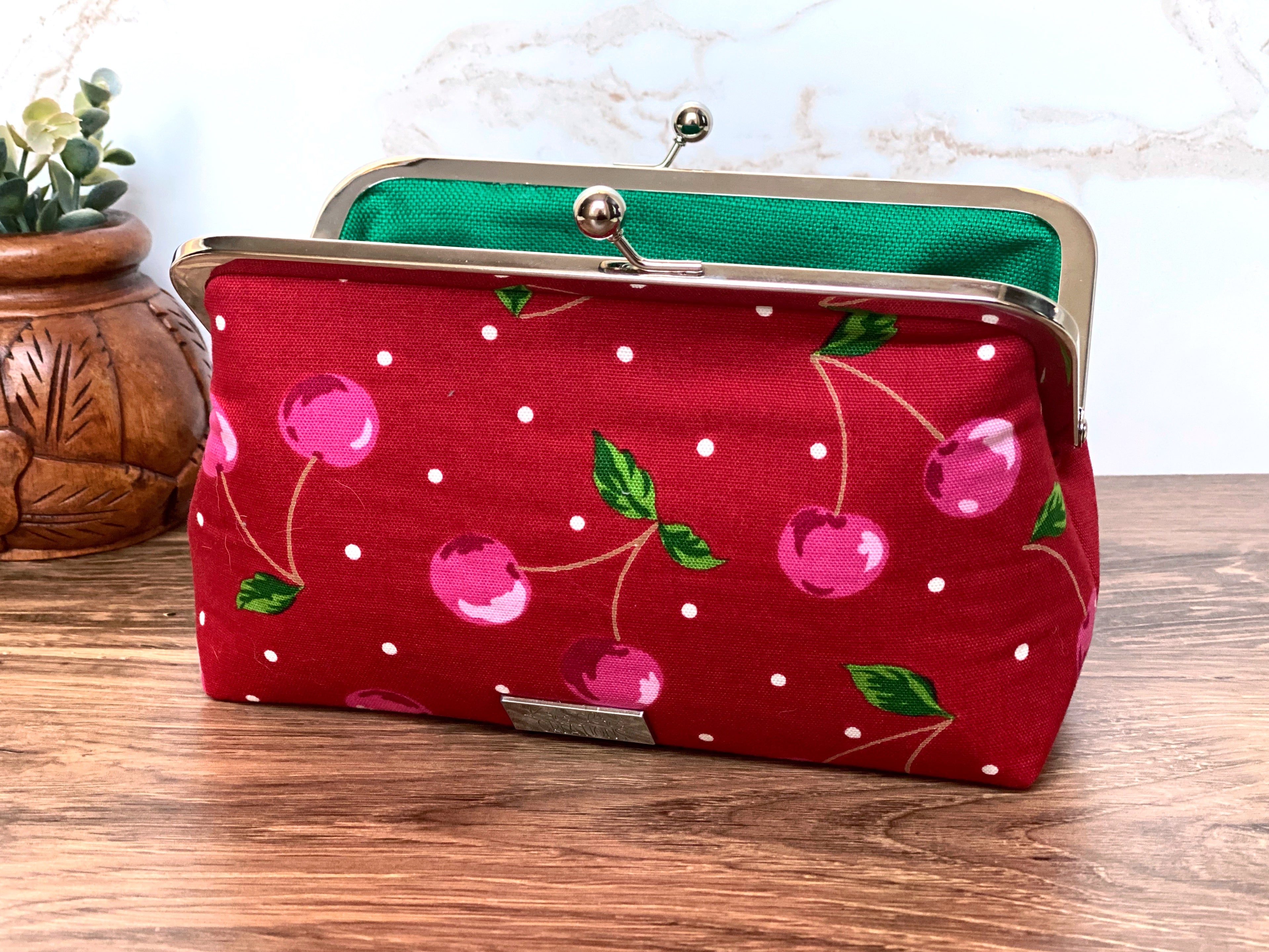 Opened twist lock clutch show green canvas lining, silver hardware, and bright cherry themed canvas exterior along with silver KrisChris tag.