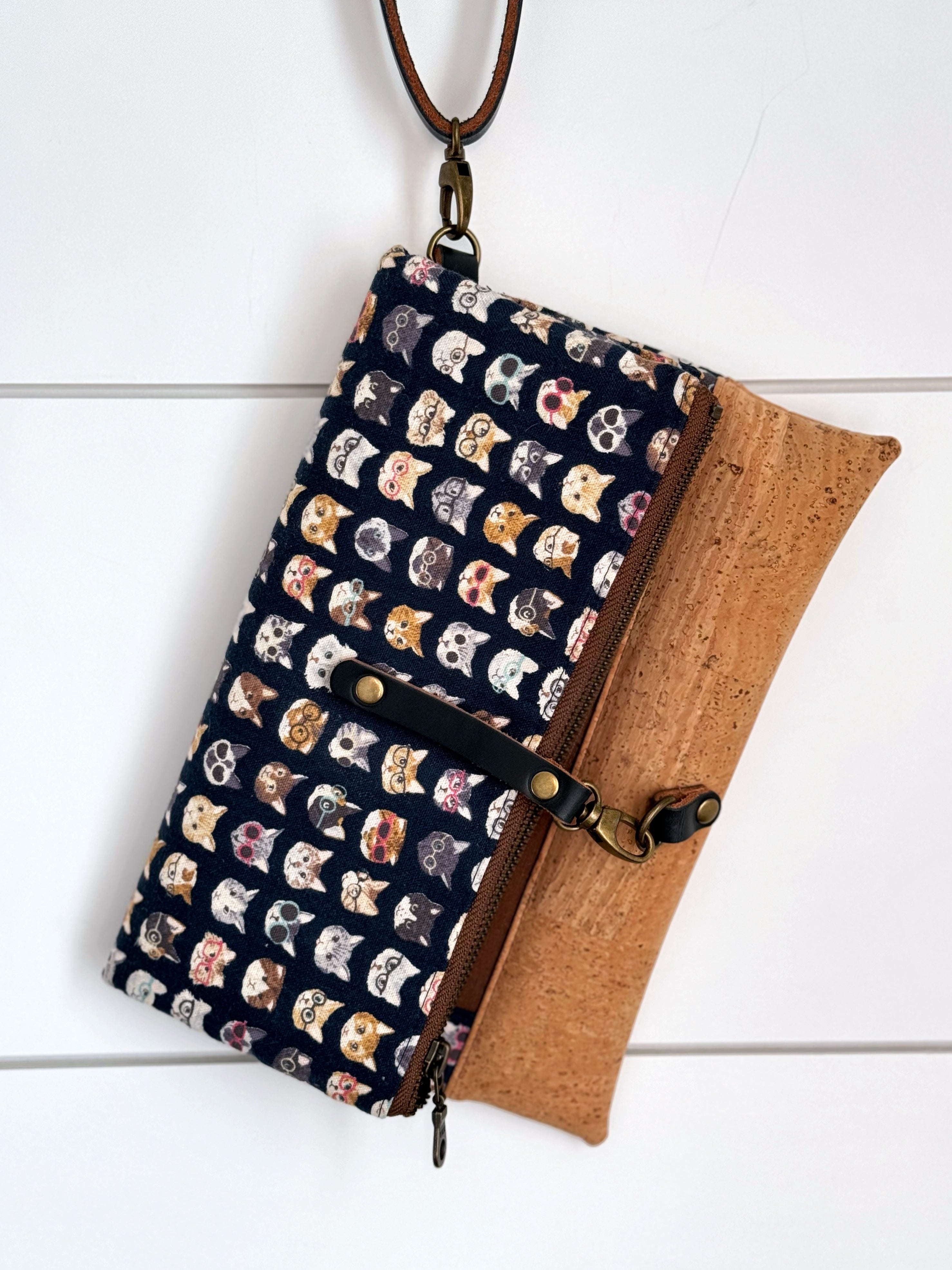 Clutch is hanging on white background from detachable leather wrist strap. Bag is clasped shut in smallest position and navy leather straps and brass rivets and lobster clasps hold it shut.