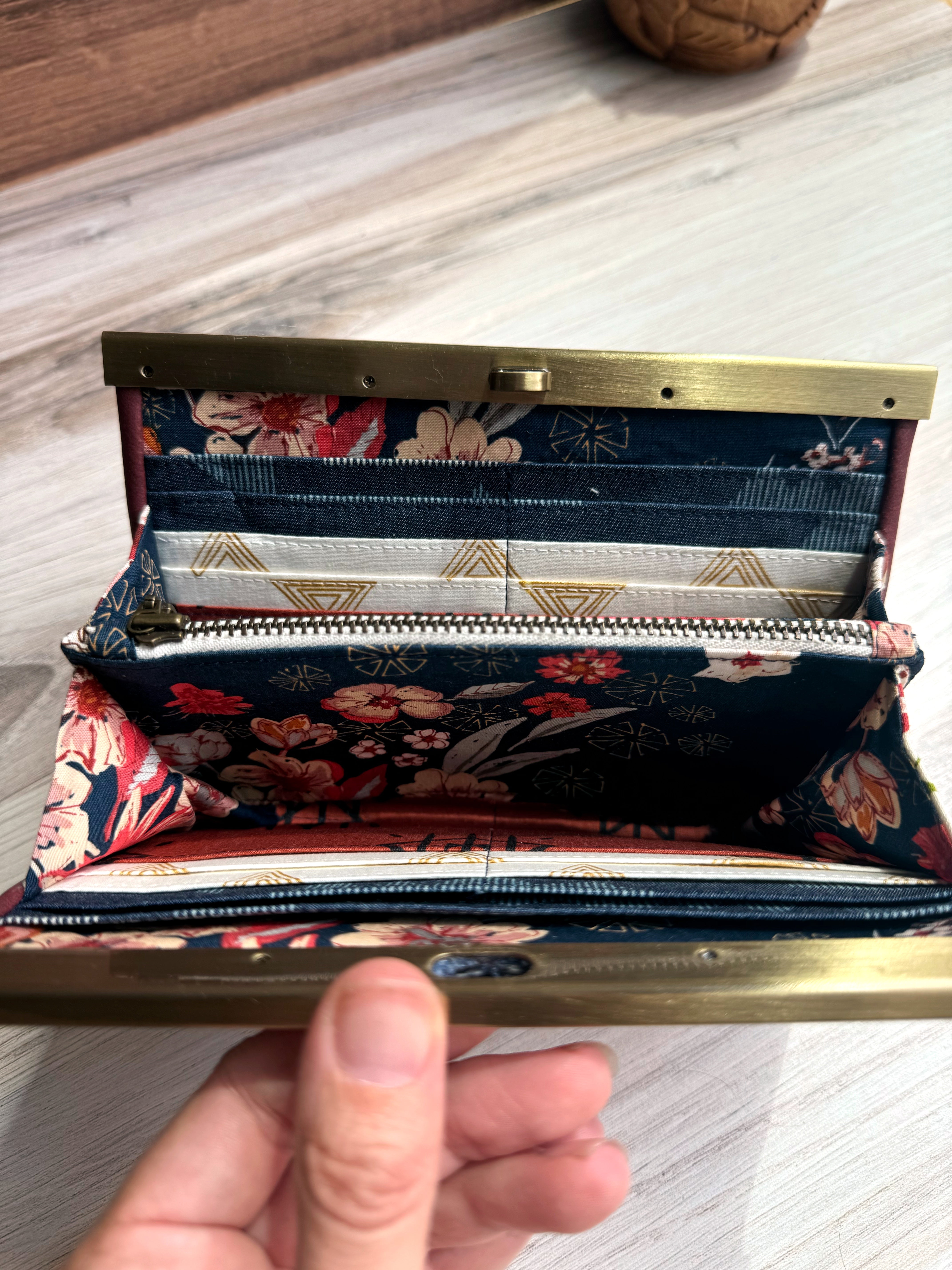 Open accordion wallet showing size scale next to a thumb. Shows detail of floral navy fabrics and geometric fabric designs on interior.