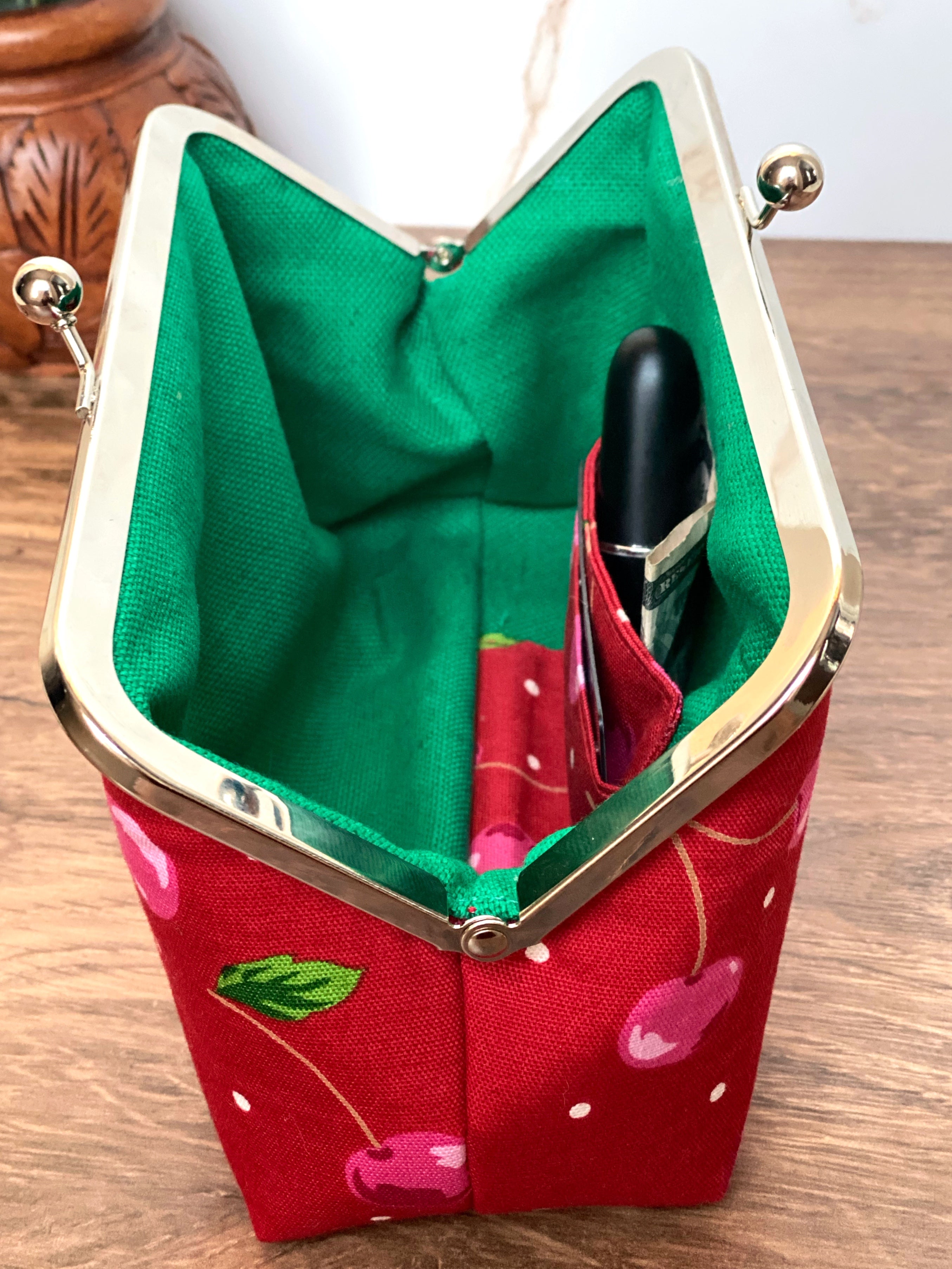 Opened kisslock clutch shows bright green canvas interior and cherry themed card and cash pockets inside along with silver twistlock hardware along the top