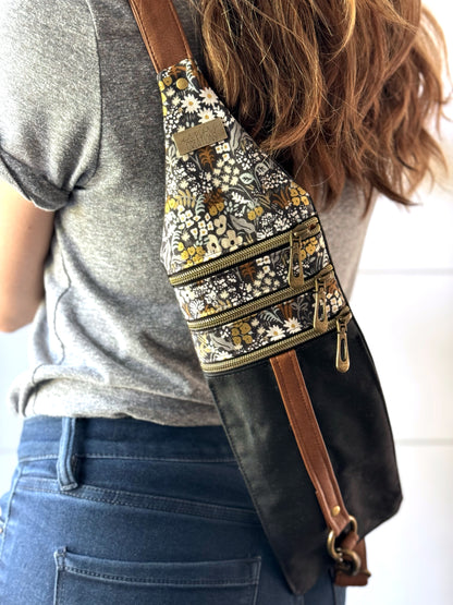 Woman in jeans wearing black and tan floral crossbody bag on the back.  Showing the 3 brass zippers across the top and adjustable vinyl strap. 