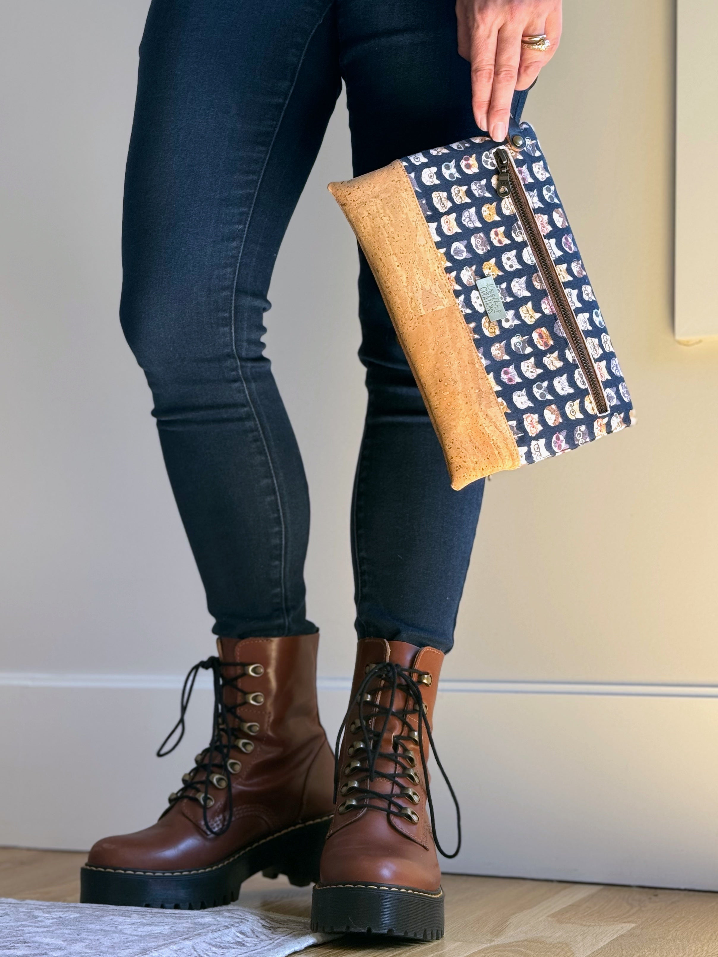 Cat clutch in woman's hand wrapped around wrist with detachable wrist strap. Standing casually with jeans and leather boots