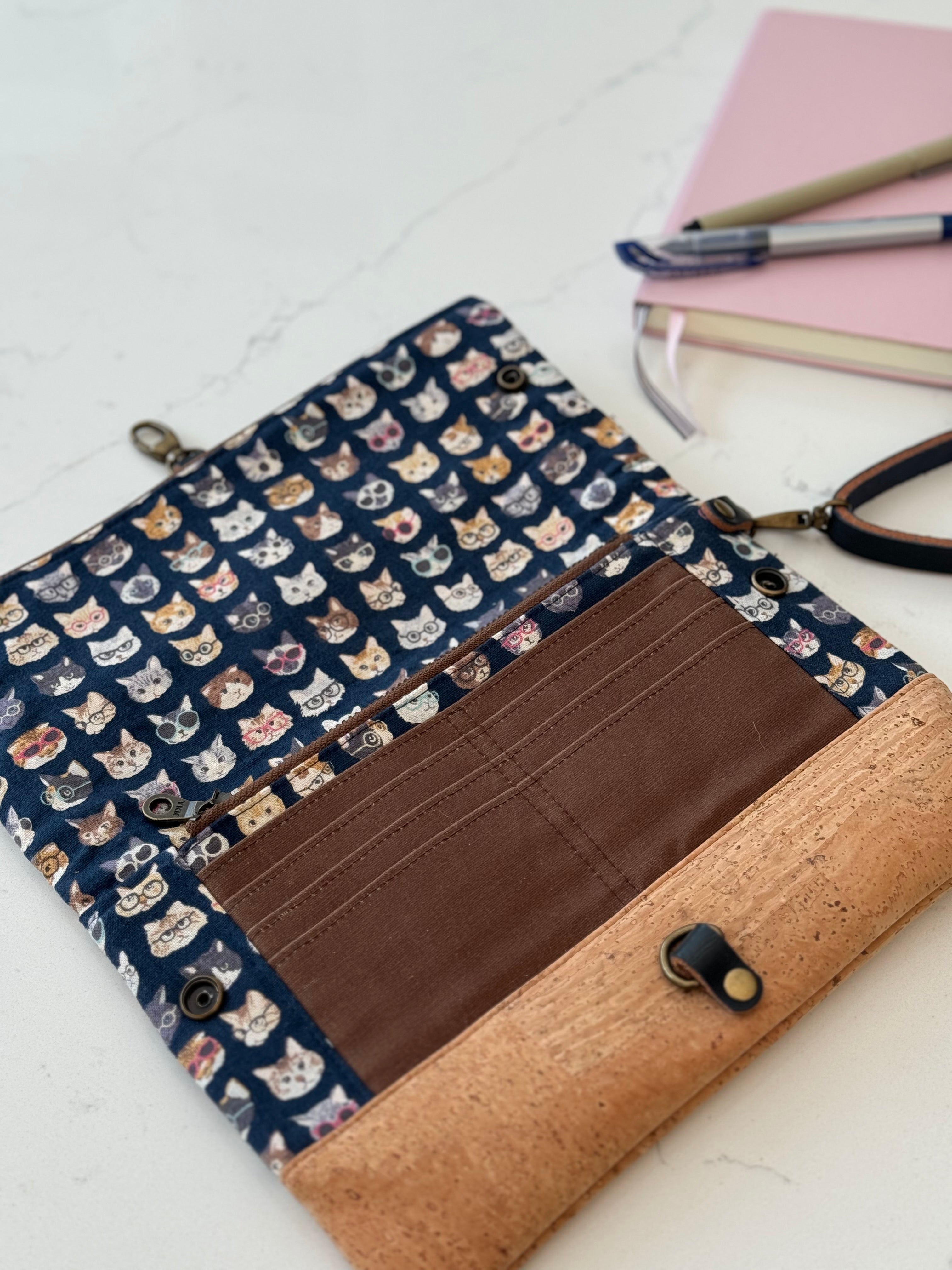Clutch bag is opened flat on table to show full size with interior zippered pocket, 8 card slots in brown waxed canvas. Tan cork base is at the bottom.