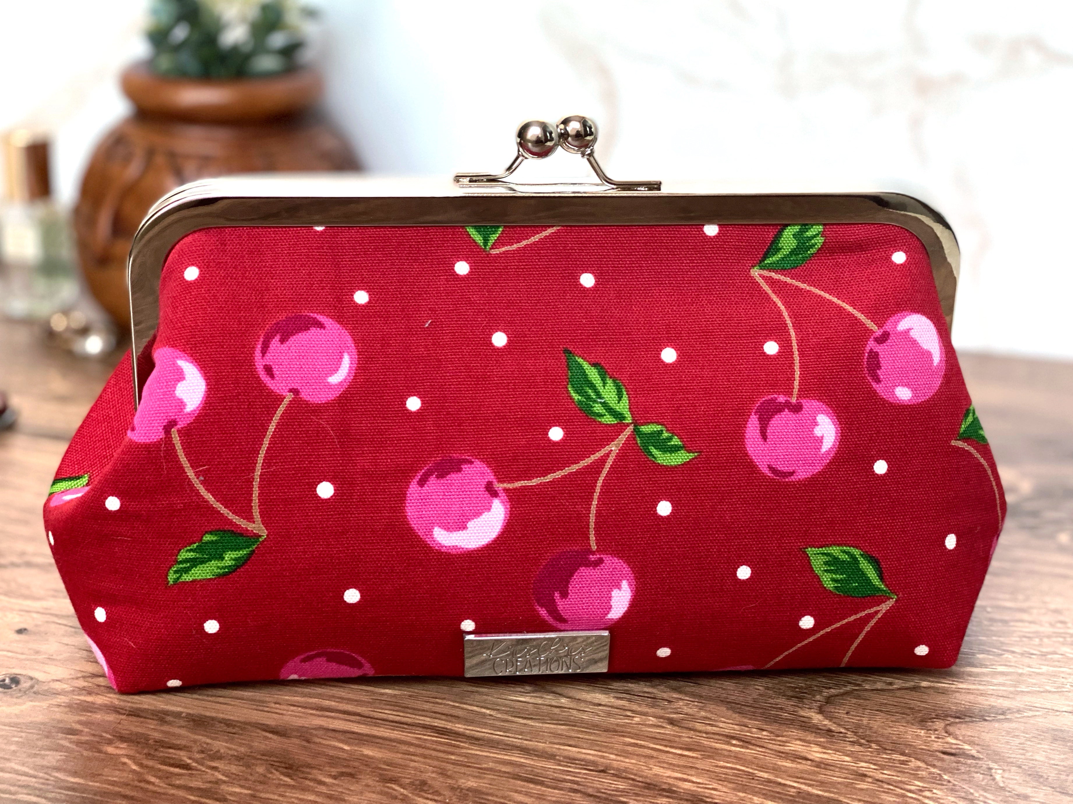 Cherry red canvas kisslock clutch shows shiny silver clasp hardware on top and bright pink cherries with green stems all over along with small white dots.