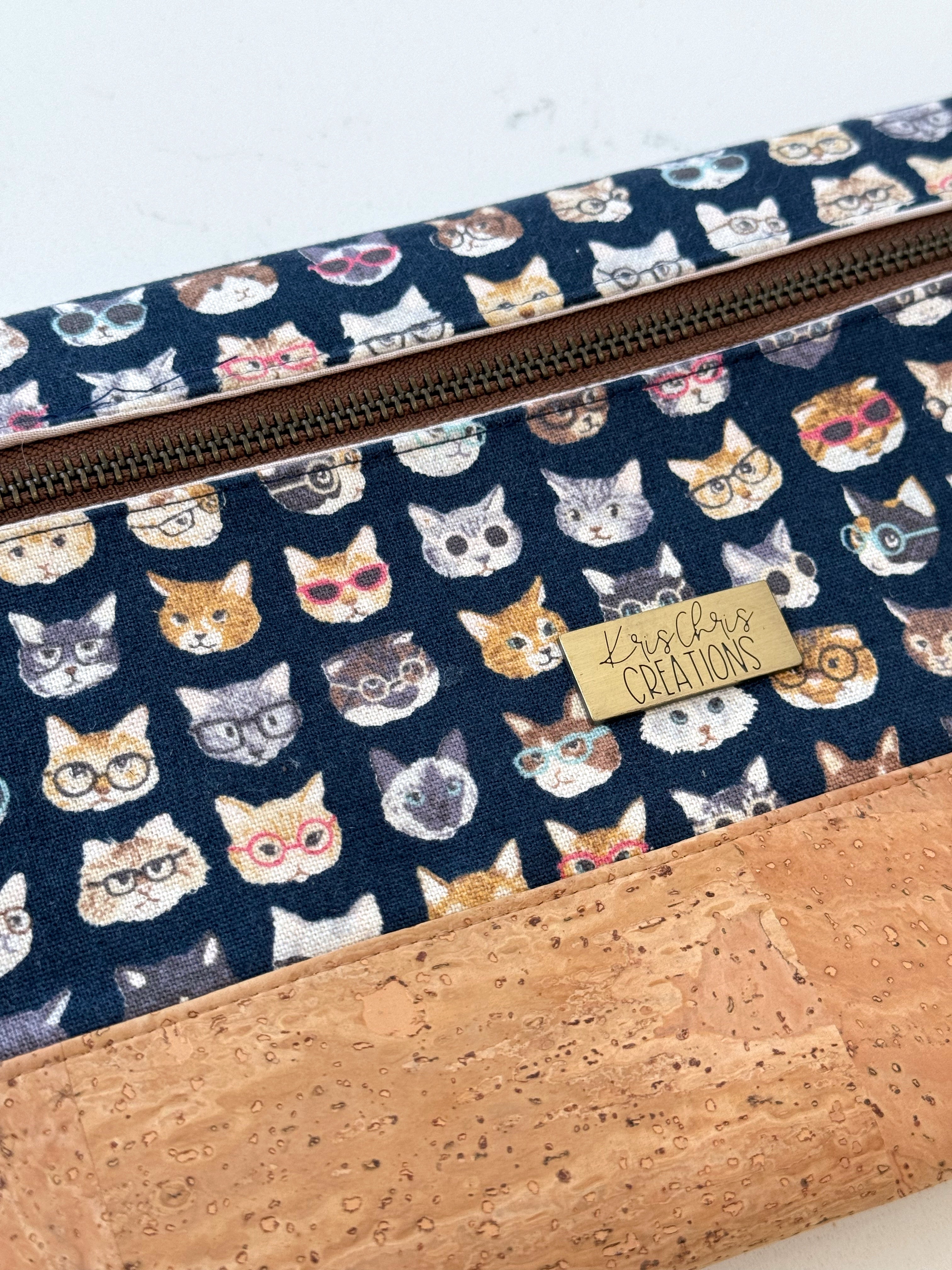 Close view of cat clutch bag shows outside zipper, variety of cats and glasses, light tan cork fabric base, and KrisChris brass label.
