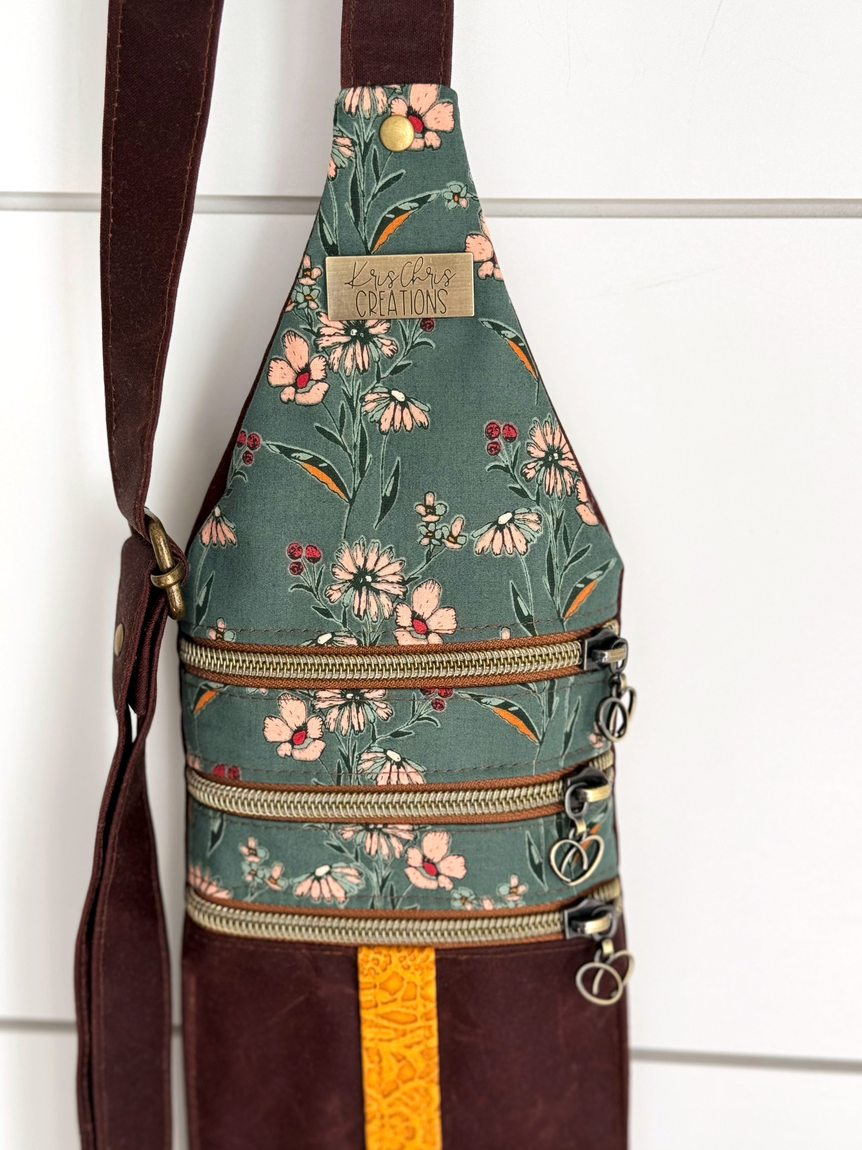 Top of teal floral bag shows adjustable strap, KrisChris brass label, brass rivet, and top 3 horizontal zippers.