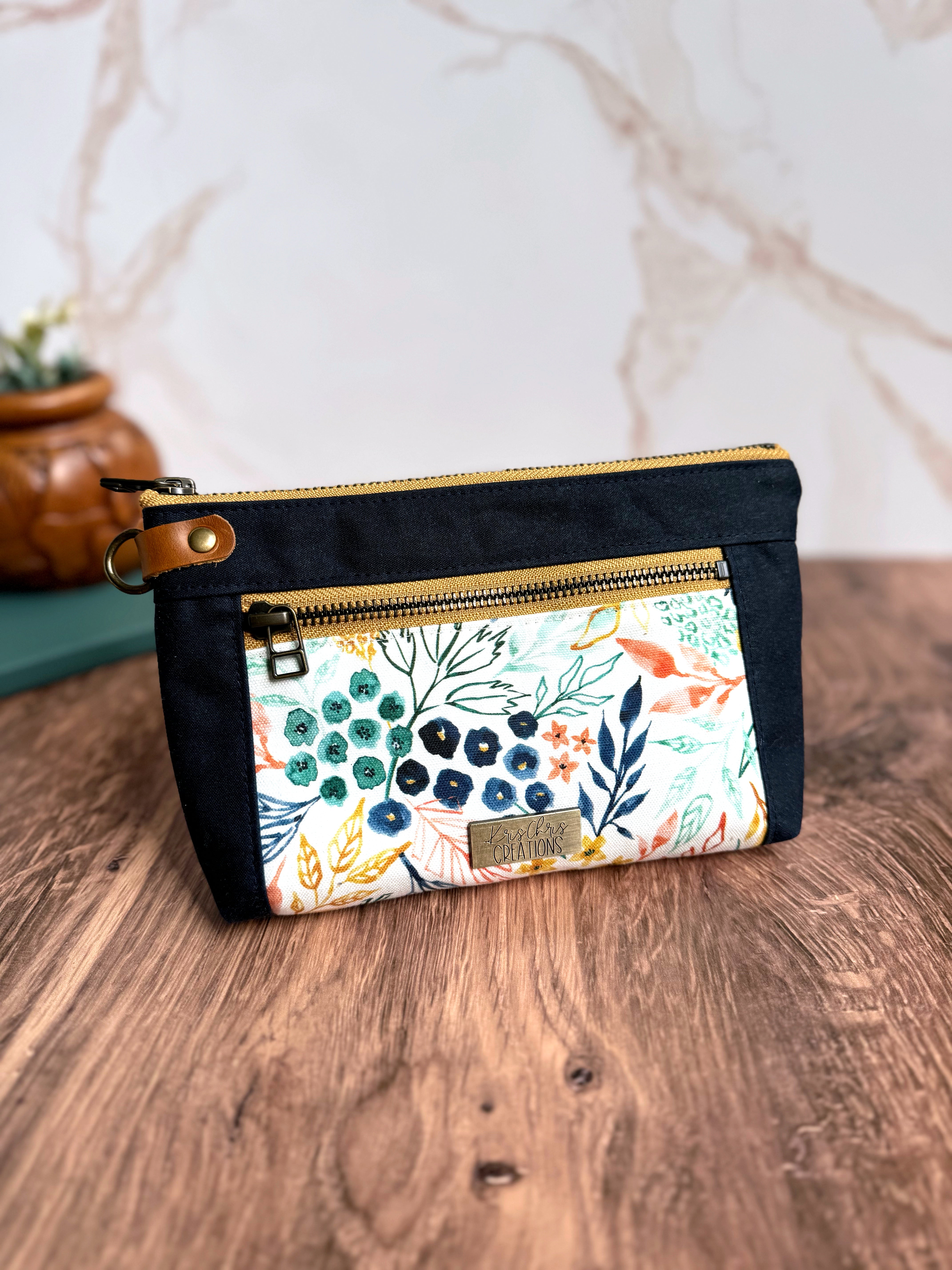 Floral pouch with Navy Waxed Canvas trim, yellow zippers, and leather tab on wooden floor and marble background.