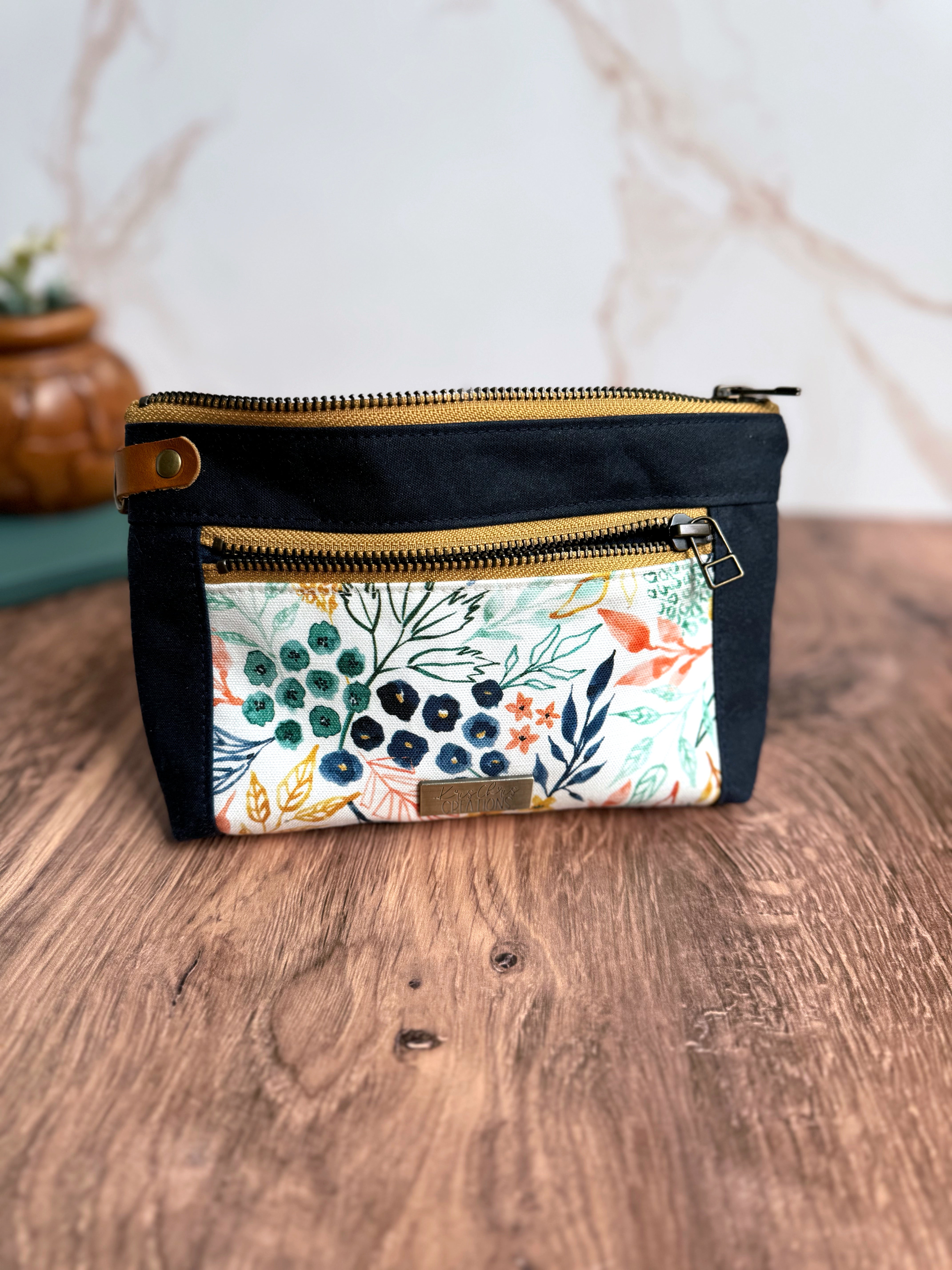 Floral pouch with Navy Waxed Canvas trim, yellow zippers, and leather tab on wooden floor and marble background.