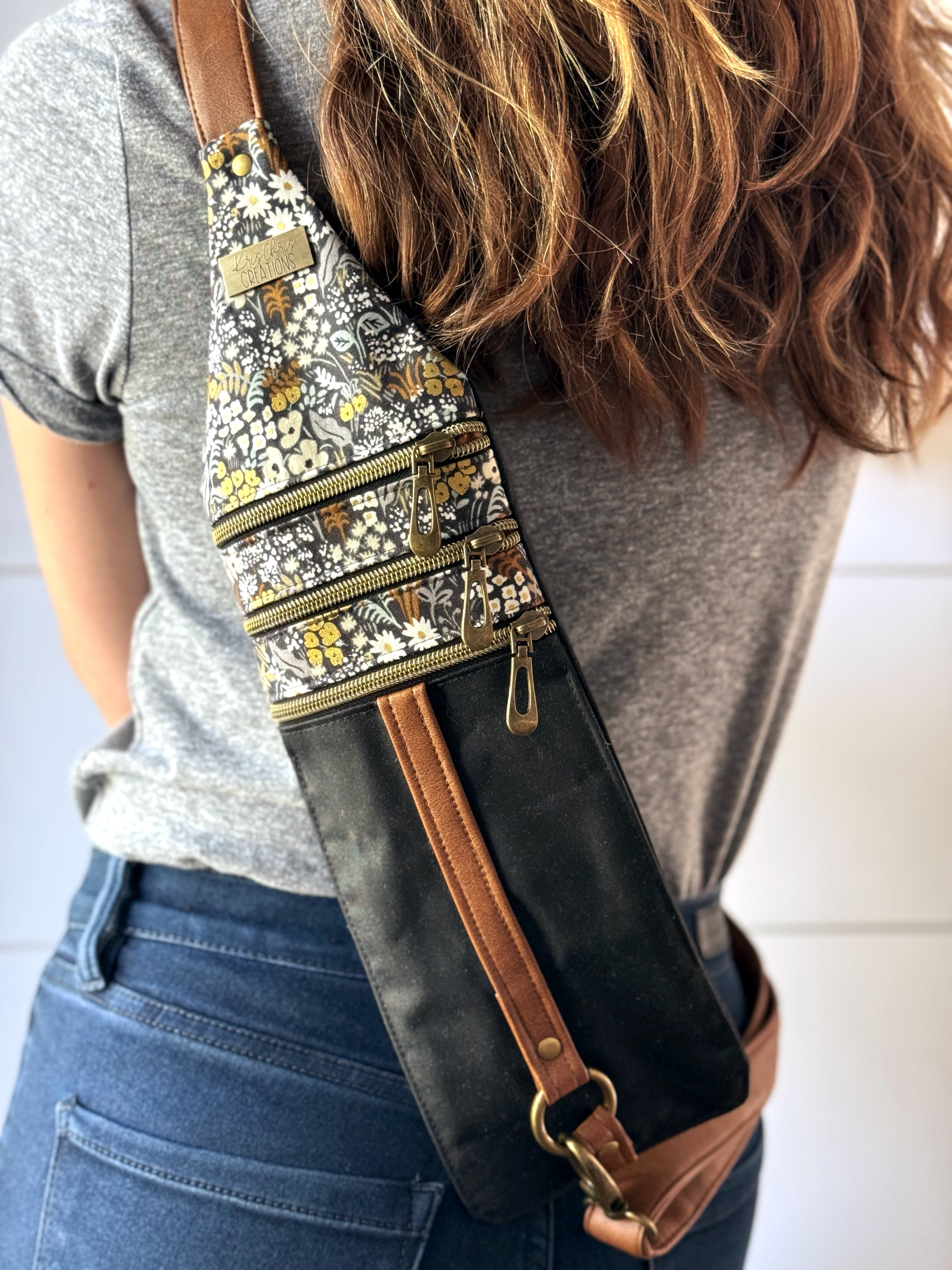 Woman wearing crossbody bag on back which has tan vegan leather straps and 3 brass zippers across. Fabric is black waxed canvas on bottom and coordinating floral fabric on top.