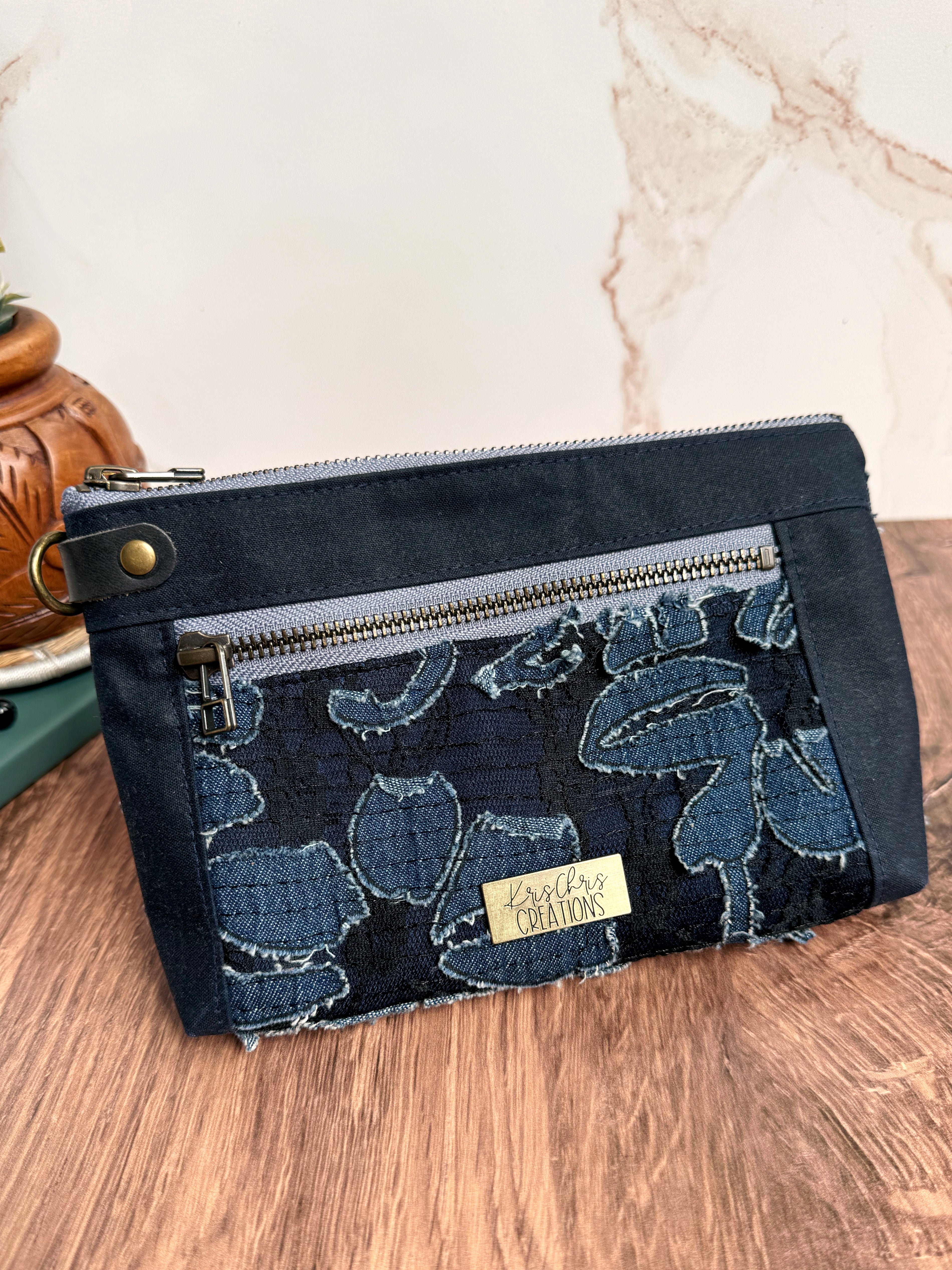 Navy pouch with solid trim, light blue zipper, and textured floral detail, and navy leather side D ring tab. 