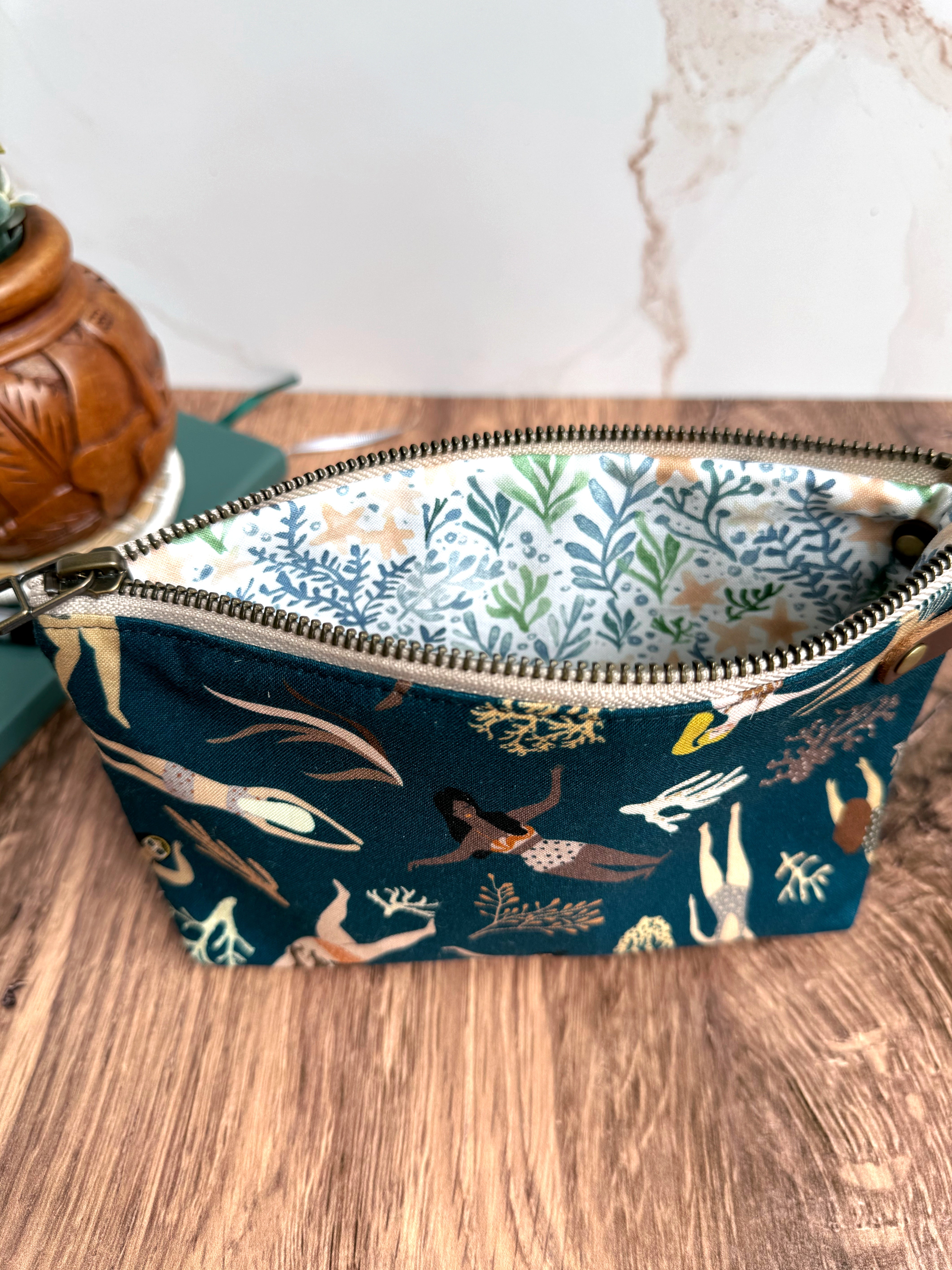 Open zipper on swimming lady pouch shows beautifully illustrated fabric on lining featuring seaweed and starfish in blue, green, and peach. 