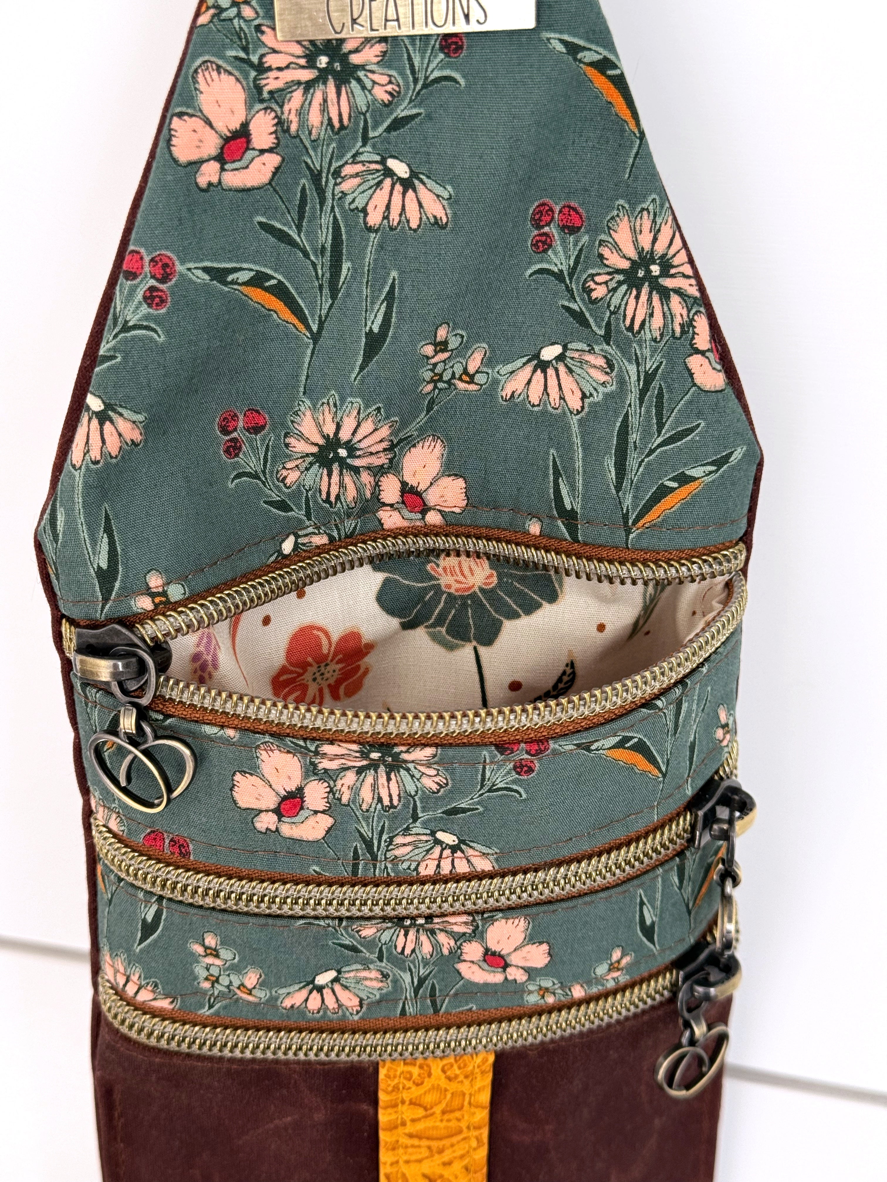 Closeup of crossbody bag with open zipper showing cream white floral fabric. Brass zippers have heart pulls. Teal Art Gallery fabrics have little flowers with pink and yellow details.