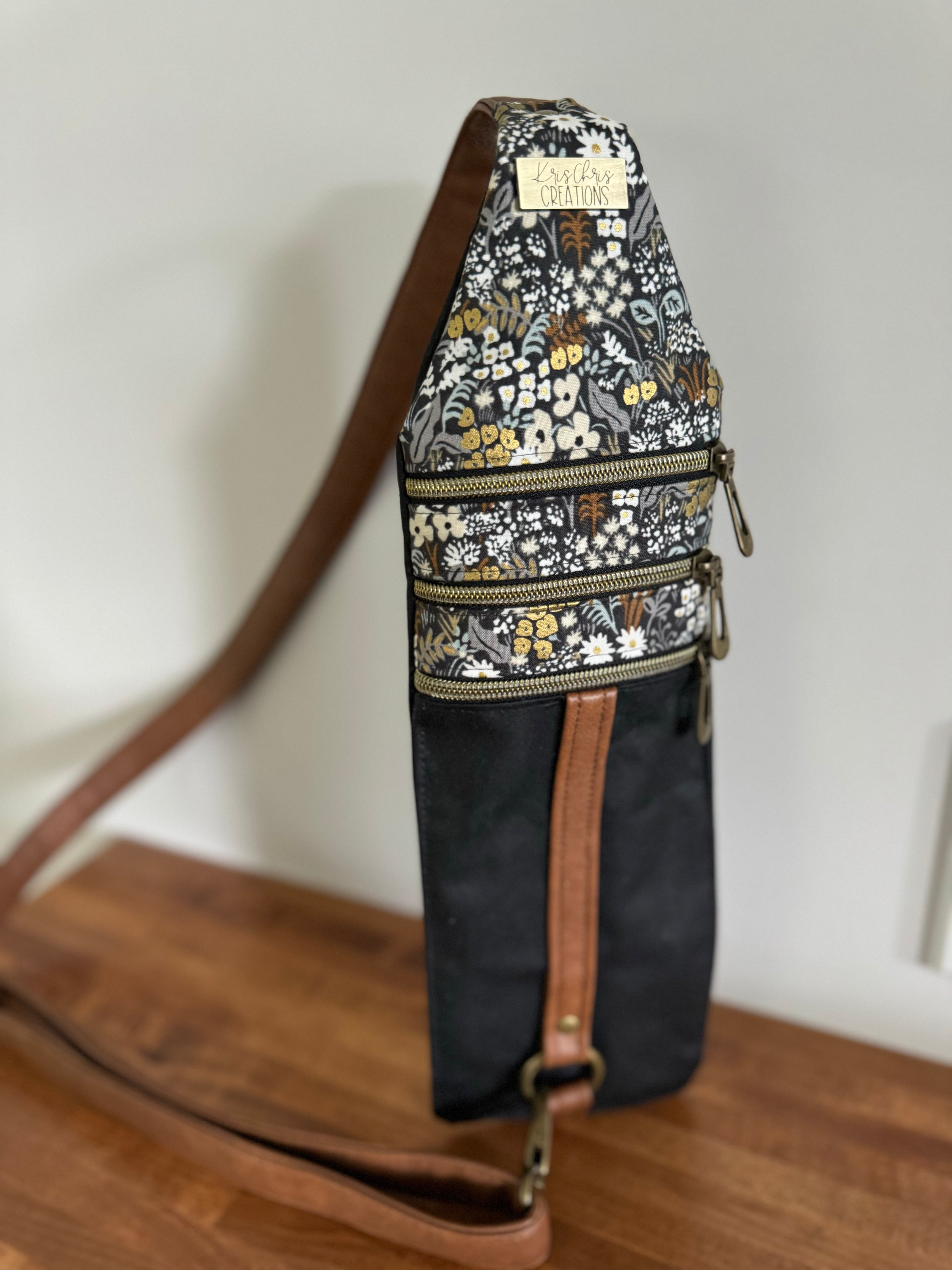 Black and tan floral crossbody bag with brass KrisChris tag.  3 zippers cross at the top and a tan vinyl strap extends vertically down to the connector across the body