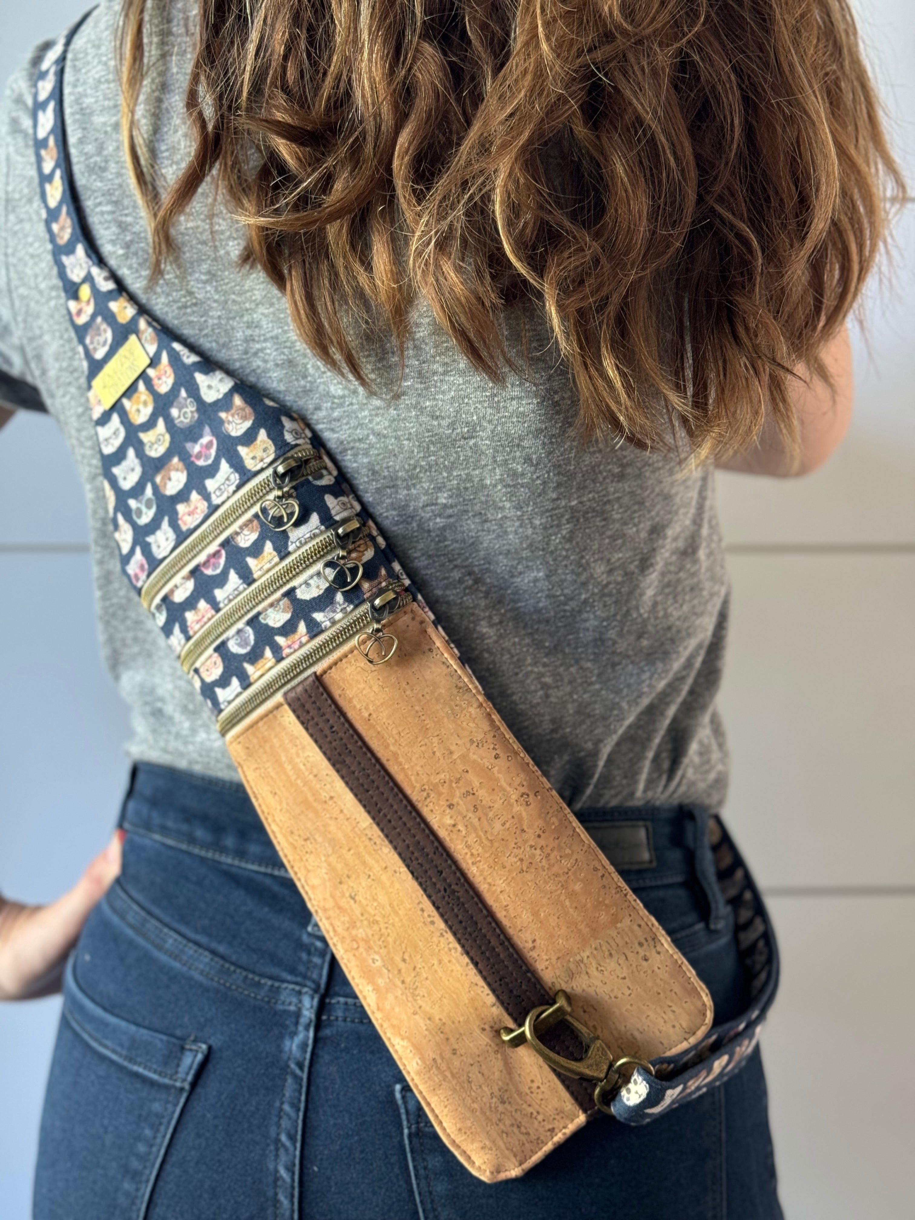 Crossbody tan cork bag with cat fabric worn on woman's back. Adjustable strap and brass hardware. 